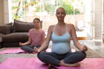 Front view of a young Caucasian pregnant woman doing yoga with her tween daughter in their sitting room — Stock Photo