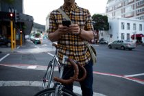 Frontansicht mittlerer Abschnitt eines jungen kaukasischen Mannes, der abends mit einem Fahrrad in einer belebten Stadtstraße steht und ein Smartphone benutzt — Stockfoto