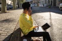 Vista lateral de um jovem adulto transexual de raça mista na moda na rua, usando um computador portátil e falando no smartphone — Fotografia de Stock