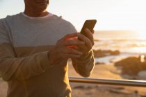 Вид спереду крупним планом зрілого Кавказького чоловіка за допомогою смартфона біля моря на заході сонця — стокове фото