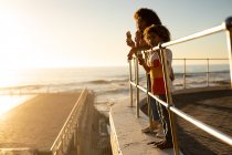 Seitenansicht einer Mischlingsfrau und ihres Sohnes im Teenageralter, die die gemeinsame Zeit am Meer genießen, die Aussicht von einer Promenade bewundern und an einem sonnigen Tag Eis essen — Stockfoto
