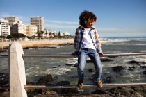 Front view of a mixed race pre-teen boy enjoying time by the sea and sitting on a balustrade on a sunny day — Stock Photo