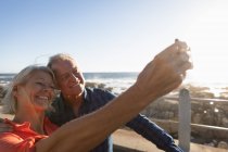 Vista lateral de cerca de un hombre y una mujer caucásicos maduros tomando una selfie junto al mar - foto de stock