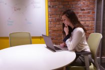 Вид сбоку на молодую кавказку, работающую в офисе креативного бизнеса, сидящую за столом с помощью ноутбука — стоковое фото