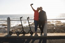 Vista frontal de un hombre y una mujer caucásicos maduros tomando una selfie al lado de e scooters junto al mar al atardecer - foto de stock