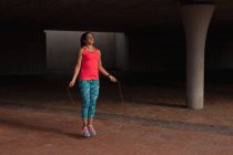 Front view of a young Caucasian woman wearing sports clothes jumping with a skipping rope during a workout under a bridge in a park — Stock Photo