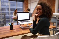 Портрет крупным планом молодой женщины смешанной расы, работающей в офисе творческого бизнеса, сидящей за столом с помощью ноутбука, поворачивающейся и улыбающейся в камеру, с коллегами, работающими за столом на заднем плане — стоковое фото