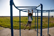 Vista frontale di un ragazzo pre-adolescente di razza mista che gioca in un parco giochi in riva al mare, appeso a una cornice di arrampicata in una giornata di sole — Foto stock