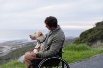 Vista lateral de um jovem caucasiano em uma cadeira de rodas passeando com seu cachorro no campo junto ao mar, acariciando o cachorro no joelho — Fotografia de Stock