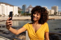 Vista frontal de una joven mestiza tomando una selfie en un día soleado junto al mar - foto de stock