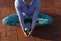 Высокий низкий вид секции женщины в спортивной одежде, сидящей на тропинке, держащей ноги и растягивающейся во время тренировки в парке — стоковое фото