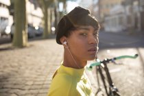 Portait de um jovem elegante mestiço transexual adulto na rua, com fones de ouvido sentados ao lado de uma bicicleta — Fotografia de Stock