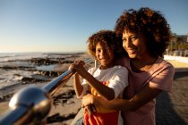 Vista frontal close-up de uma mulher de raça mista e seu filho pré-adolescente desfrutando de tempo juntos junto ao mar, abraçando e sorrindo em um dia ensolarado — Fotografia de Stock