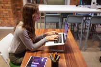 Vue de côté gros plan d'une jeune femme caucasienne travaillant dans le bureau d'une entreprise créative assis à un bureau à l'aide d'un ordinateur portable — Photo de stock