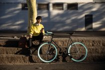 Vue de face d'un jeune transgenre de race mixte à la mode adulte dans la rue, tenant un smartphone et une tasse de café assis sur des marches à côté d'un vélo — Photo de stock