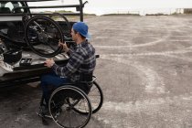 Вид збоку молодого Кавказького чоловіка в інвалідному візку приймаючи лежачий велосипед з задньої частини своєї машини, щоб зібрати його на автостоянці біля моря — стокове фото