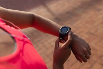 Над пліч-о-пліч жінка носить спортивний одяг, перевіряючи свій розумний годинник і слухаючи музику на навушниках, працюючи сонячно в парку — стокове фото