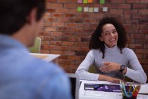 За плечами молодой женщины смешанной расы и молодого кавказского коллеги, разговаривающего в офисе творческого бизнеса, женщина улыбается и указывает — стоковое фото