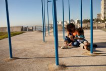 Vista frontal de una joven mestiza y su hijo preadolescente disfrutando del tiempo juntos jugando en un parque infantil junto al mar, sentados en el suelo por un marco de escalada y tomando selfies en un día soleado - foto de stock