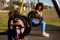 Вид спереди на молодую расовую женщину и ее сына-подростка, наслаждающуюся совместной игрой на детской площадке, фотографирующуюся на качелях в солнечный день — стоковое фото