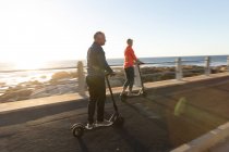 Side view of a mature Caucasian man and woman riding e scooters by the sea at sunset — Stock Photo
