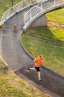 Vista lateral de un joven atlético caucásico ejercitándose en una pasarela de una ciudad, corriendo y escuchando música con auriculares - foto de stock