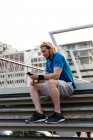 Side view of a young athletic Caucasian man exercising on a footbridge in a city, sitting on the steps and using a smartphone during a break — Stock Photo