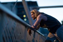 Side view of a young athletic Caucasian man exercising in a city park in the evening, resting on a handrail during a break with defocused lights in the background — Stock Photo
