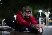Vista frontal de un joven atlético caucásico ejercitándose en un parque de la ciudad por la noche, estirándose en un banco con un faro encendido - foto de stock