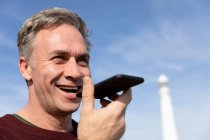 Vista frontal close-up de um homem caucasiano de meia-idade falando em um smartphone enquanto desfruta de tempo livre relaxando em uma praia perto de um farol em um dia ensolarado — Fotografia de Stock