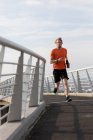 Front view of a young athletic Caucasian man exercising on a footbridge in a city, running with earphones on and holding a bottle — Stock Photo