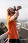 Side view of a young athletic Caucasian man exercising on a footbridge in a city, listening to music with earphones on drinking water during a break — Stock Photo
