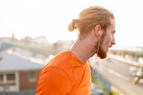 Vista lateral de un joven caucásico ejercitándose en una pasarela de una ciudad, escuchando música con auriculares durante un descanso - foto de stock