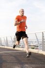 Front view of a young athletic Caucasian man exercising on a footbridge in a city, running and listening to music with earphones on — Stock Photo