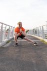 Vue de face d'un jeune homme athlétique caucasien faisant de l'exercice sur une passerelle dans une ville, étirant et écoutant de la musique avec des écouteurs allumés — Photo de stock