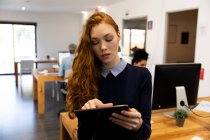 Передній вид на молоду кавказьку жінку, що працює в творчому кабінеті, стоїть за столом, тримає планшетний комп 