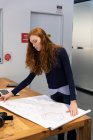 Бічний вид на молоду кавказьку жінку, що працює у творчому кабінеті, стоїть за столом, дивлячись на плани архітекторів. — стокове фото