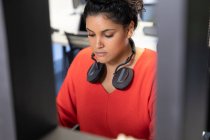 Front view of a young mixed race woman sitting at a desk in a creative office, with headphones around her neck. — Stock Photo