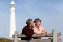 Front view of an adult Caucasian couple enjoying free time sitting on a bench and smiling near a lighthouse on a sunny day — Stock Photo