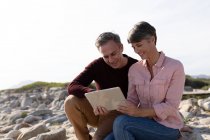 Front view of an adult Caucasian couple enjoying free time relaxing together on a beach using a tablet on a sunny day — Stock Photo