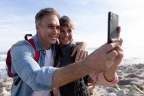 Вид спереди на кавказскую пару, использующую смартфон, наслаждающуюся свободным временем у моря на пляже в солнечный день — стоковое фото