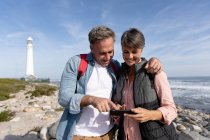 Vue de face d'un couple adulte caucasien profitant de temps libre à l'aide d'un smartphone et souriant près d'un phare au bord de la mer par une journée ensoleillée — Photo de stock