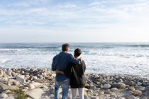 Visão traseira de um casal adulto caucasiano desfrutando de tempo livre abraçando juntos em uma praia ao lado do mar em um dia ensolarado — Fotografia de Stock