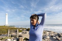 Вид спереди на кавказскую женщину, наслаждающуюся свободным временем, растягиваясь перед упражнениями у маяка на берегу моря в солнечный день — стоковое фото