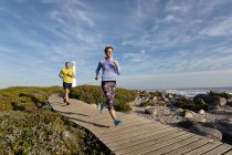 Front view of an adult Caucasian couple enjoying free time running a path near a lighthouse beside the sea on a sunny day — Stock Photo