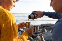 Vista frontale primo piano di una coppia caucasica adulta che si gode il tempo libero rilassandosi insieme su una spiaggia vicino al mare bevendo caffè in una giornata di sole — Foto stock