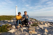 Вид спереди на взрослую кавказскую пару, наслаждающуюся свободным временем, отдыхающую вместе на пляже у моря, пьющую кофе возле маяка в солнечный день — стоковое фото