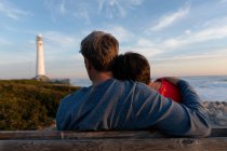 Задний план вид на взрослую кавказскую пару, наслаждающуюся свободным временем, сидя на скамейке и обнимаясь рядом с морем возле маяка в солнечный день — стоковое фото