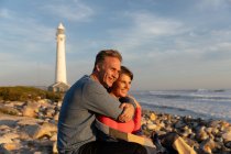 Vorderansicht eines erwachsenen kaukasischen Paares, das an einem sonnigen Tag in der Nähe eines Leuchtturms die freie Zeit genießt und sich gemeinsam am Strand umarmt — Stockfoto