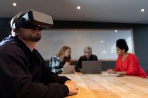 Vista laterale di un giovane caucasico seduto a un tavolo in una sala conferenze creativa dell'ufficio, che indossa un visore VR con i suoi colleghi in una riunione sullo sfondo . — Foto stock